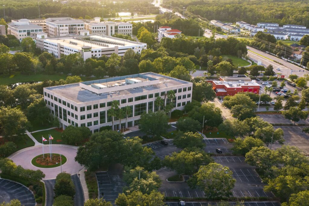 A four-story office building surrounded by trees with a large parking lot that is partially filled with cars, seen from above during daylight.