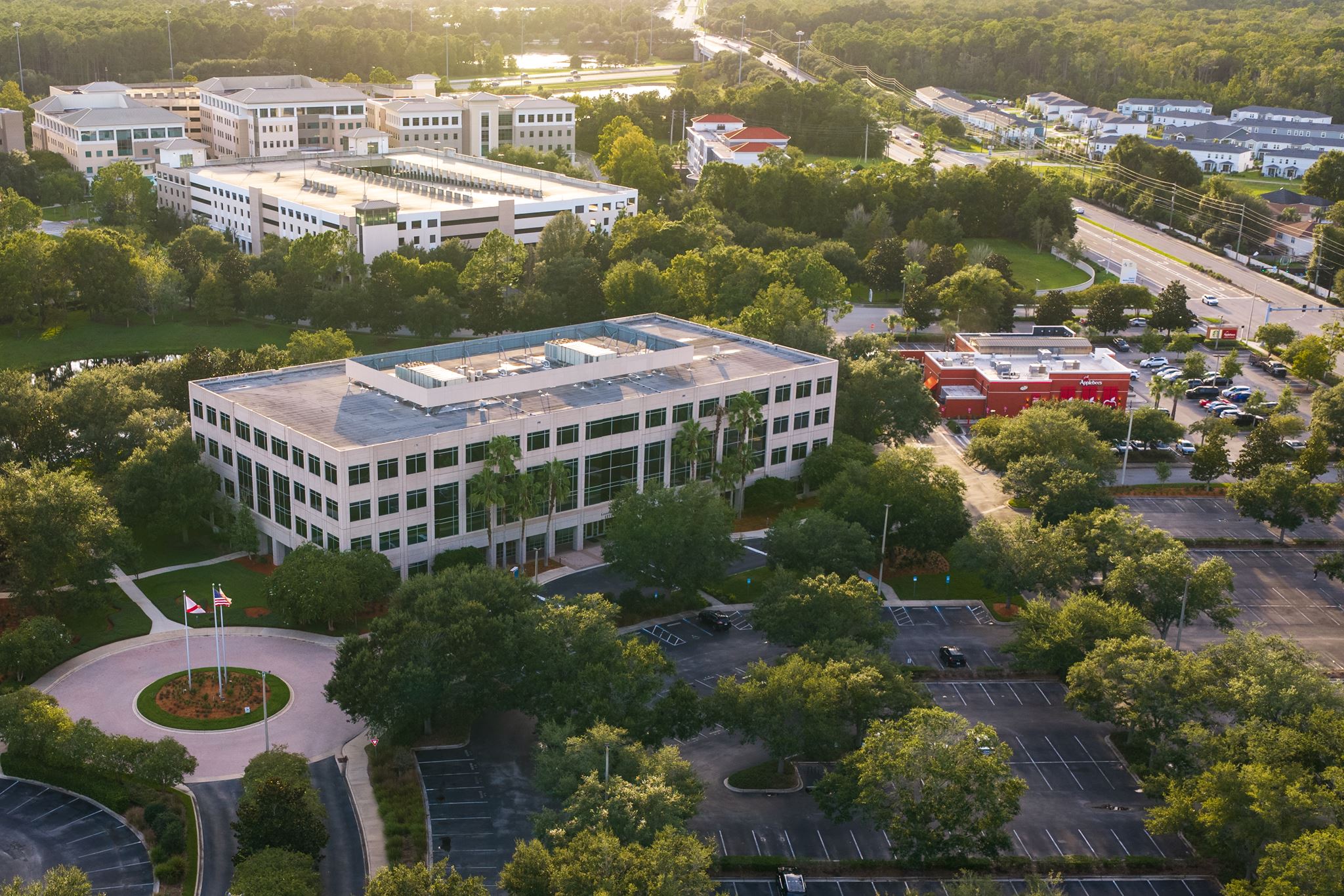 A four-story office building surrounded by trees with a large parking lot that is partially filled with cars, seen from above during daylight.