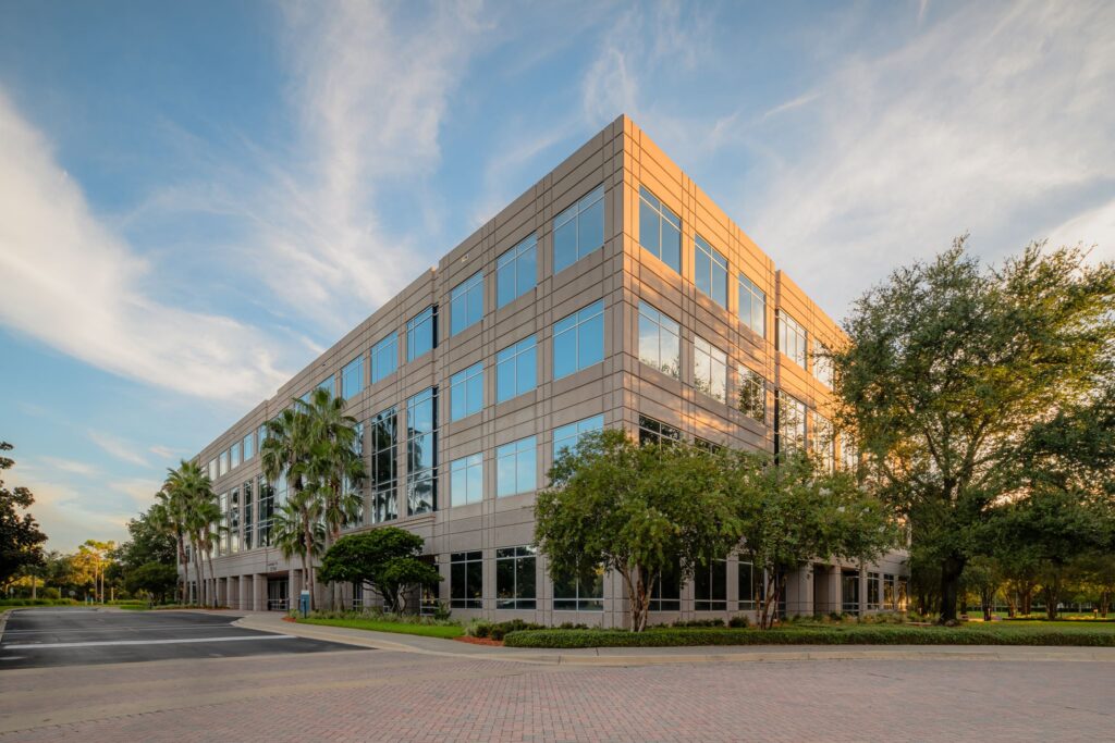 Four-story modern office building with large windows, palm trees, and surrounding greenery under a blue sky with clouds.