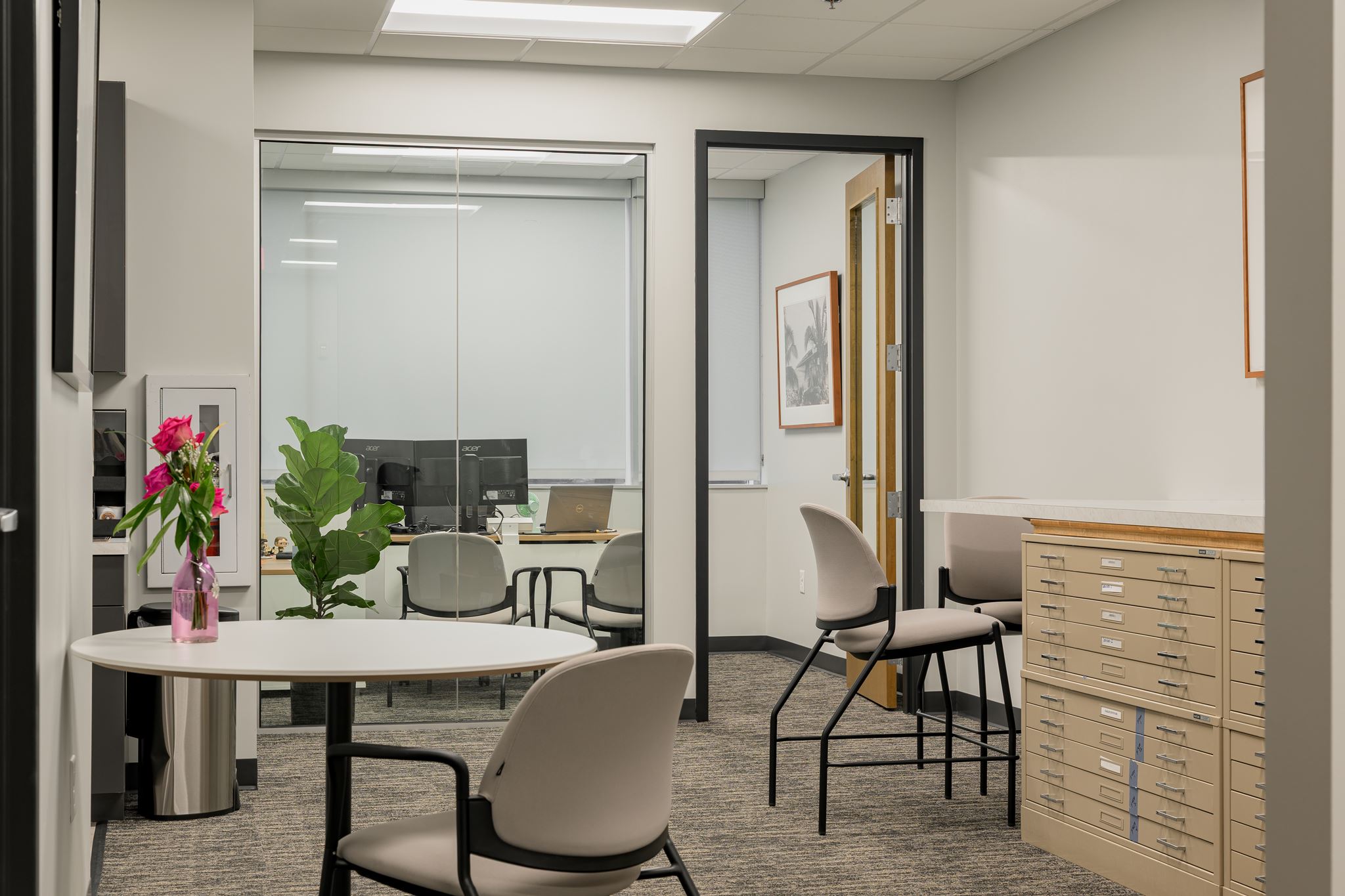 Modern office space with neutral decor, two desks, chairs, a potted plant, drawers, and a vase with flowers; glass wall reveals another office area with computer monitors.