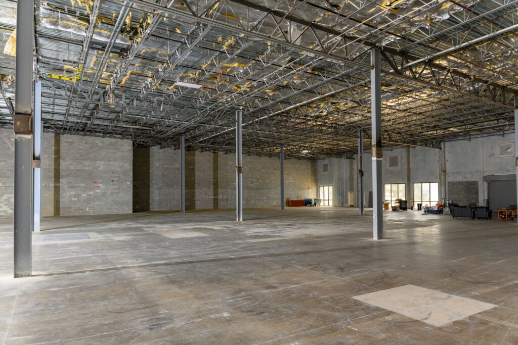 Large empty warehouse with exposed ceiling pipes and concrete walls, partially finished floors, and scattered construction equipment along the back wall.