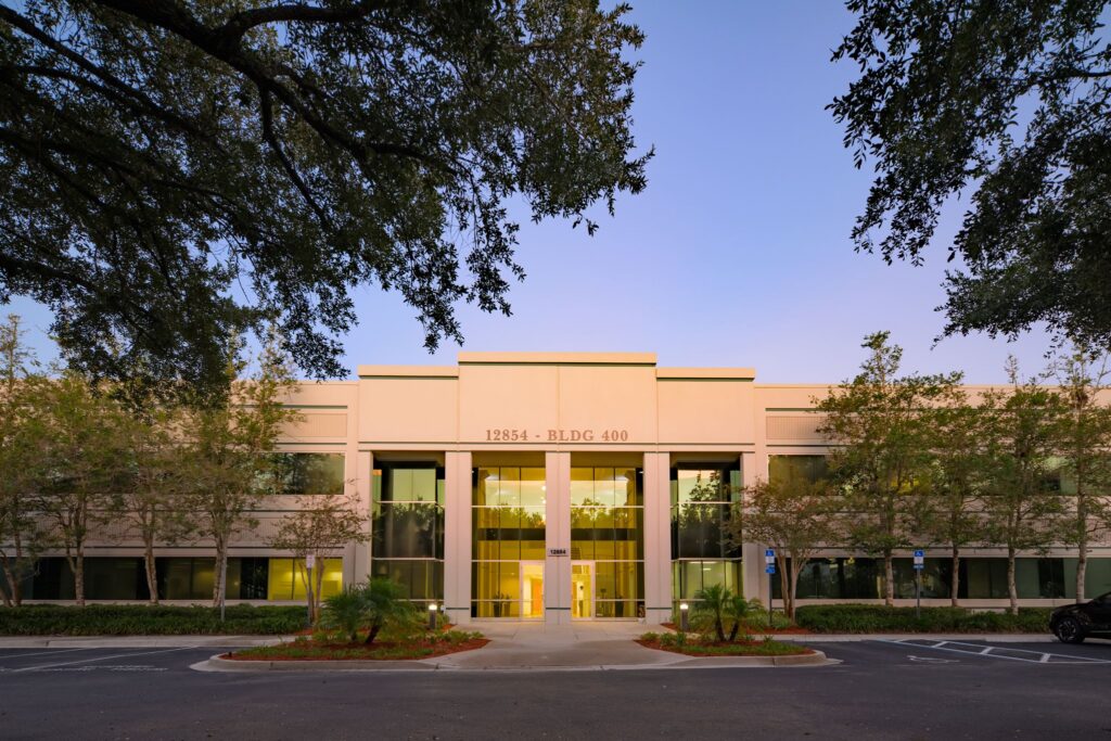 Two-story office building labeled “12854 - BLDG 400” with large windows, a central entrance, trees, and an empty parking lot in front.