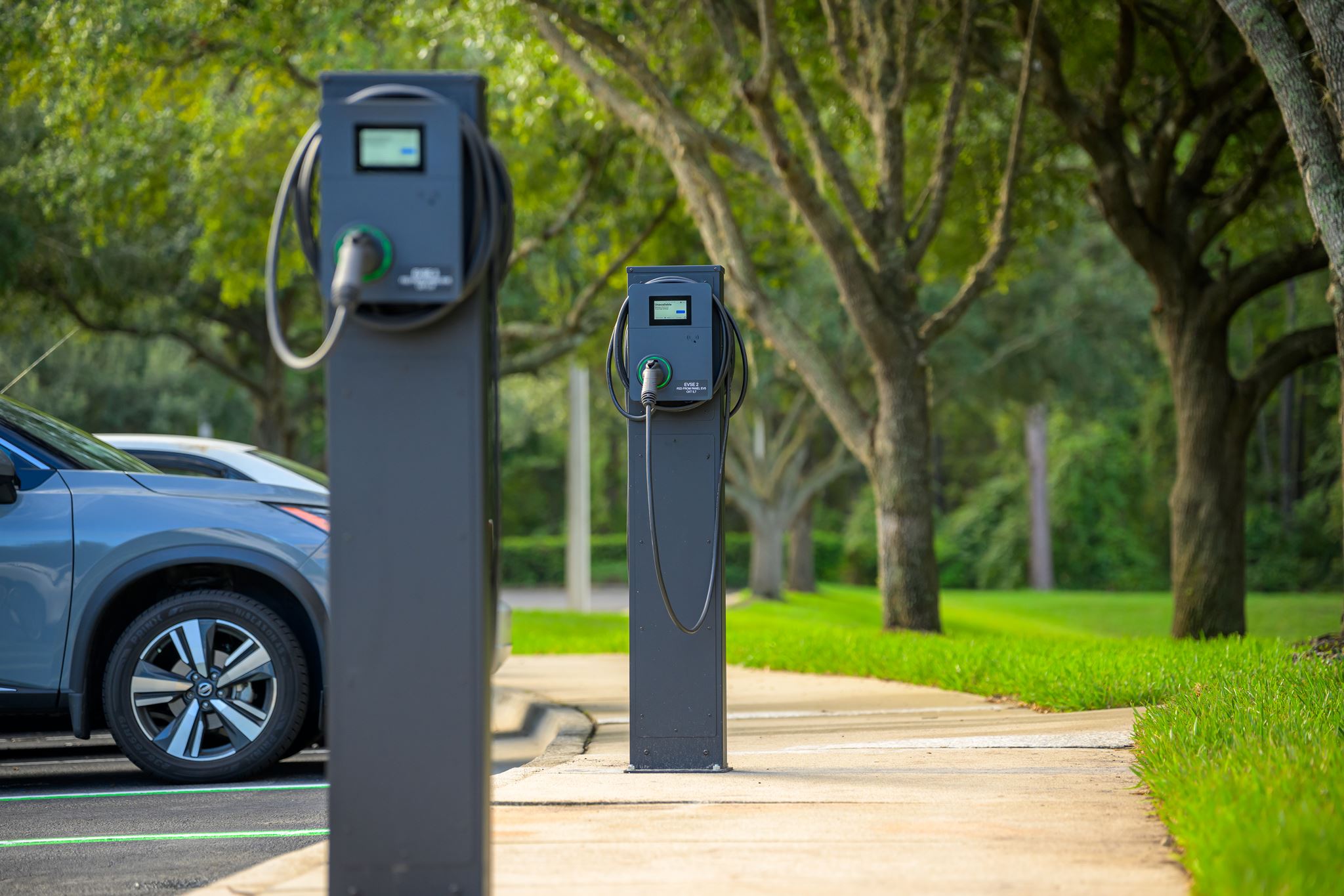 Three electric vehicle charging stations are installed along a sidewalk in a parking lot, with trees and a parked car in the background.