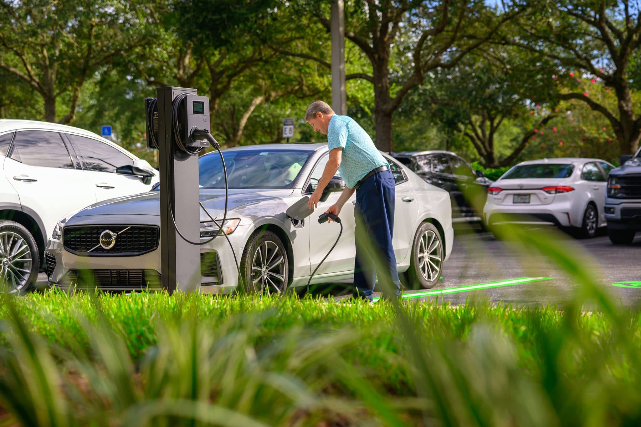 A man plugs a charging cable into a silver Volvo electric vehicle at a charging station in a parking lot with other parked cars and trees in the background.