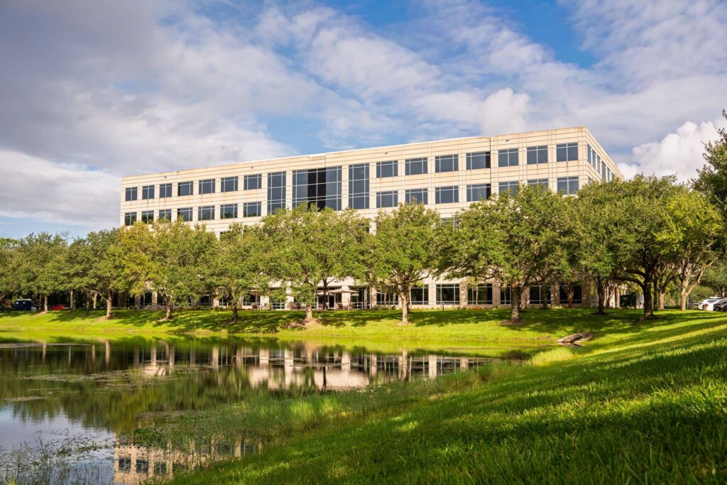 Four-story office building with large windows, surrounded by trees and reflected in a pond, under a partly cloudy sky.