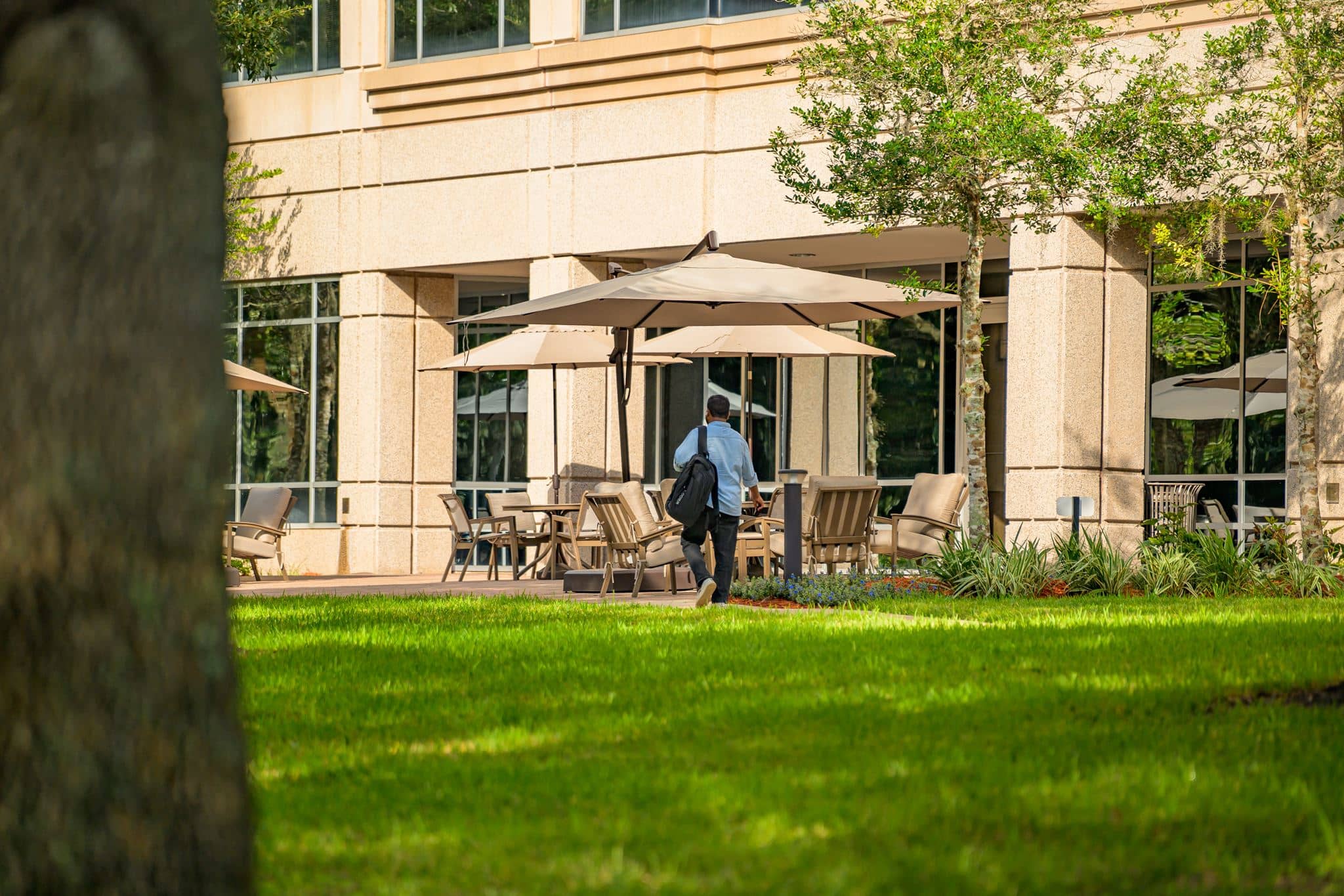 Person walking toward an outdoor seating area with umbrellas and chairs in front of a modern office building, surrounded by green grass and trees.