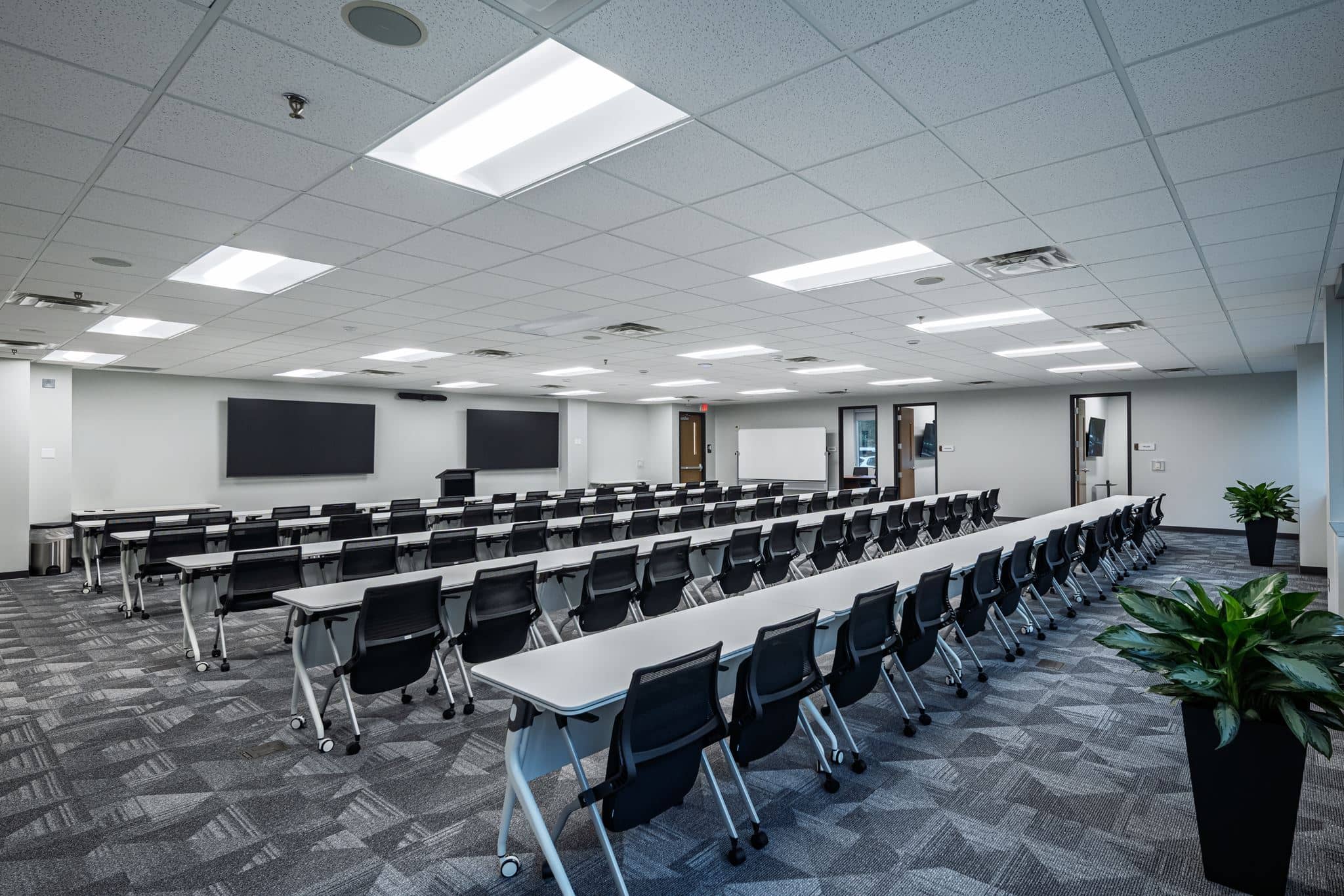 A modern, empty conference room with rows of black chairs and white tables facing several large screens and a whiteboard; two potted plants are in the corners.