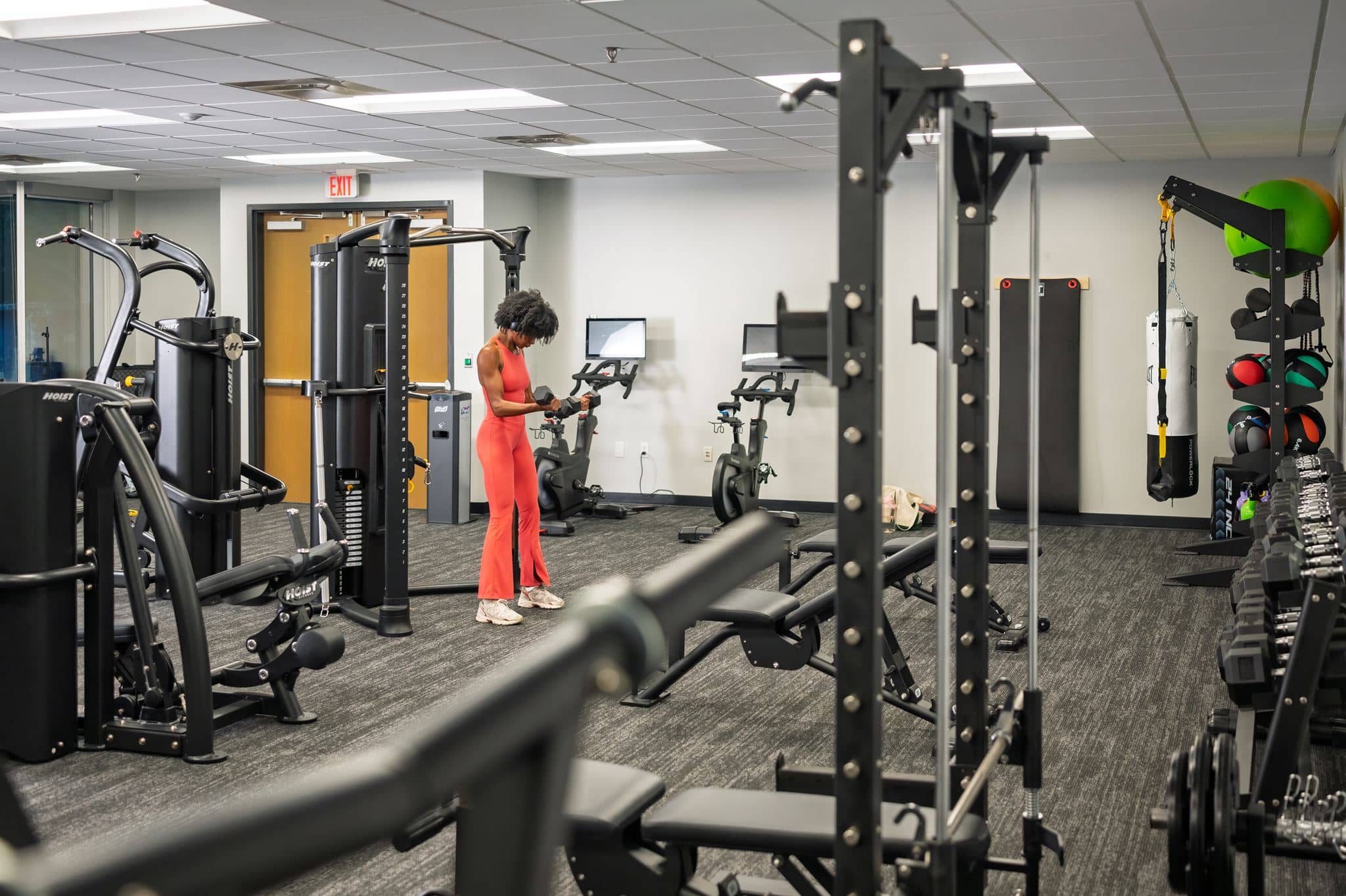A gym with dumbbells on racks, a weight bench, and exercise machines. A person in red workout clothes is using equipment near the mirrored wall in the background.