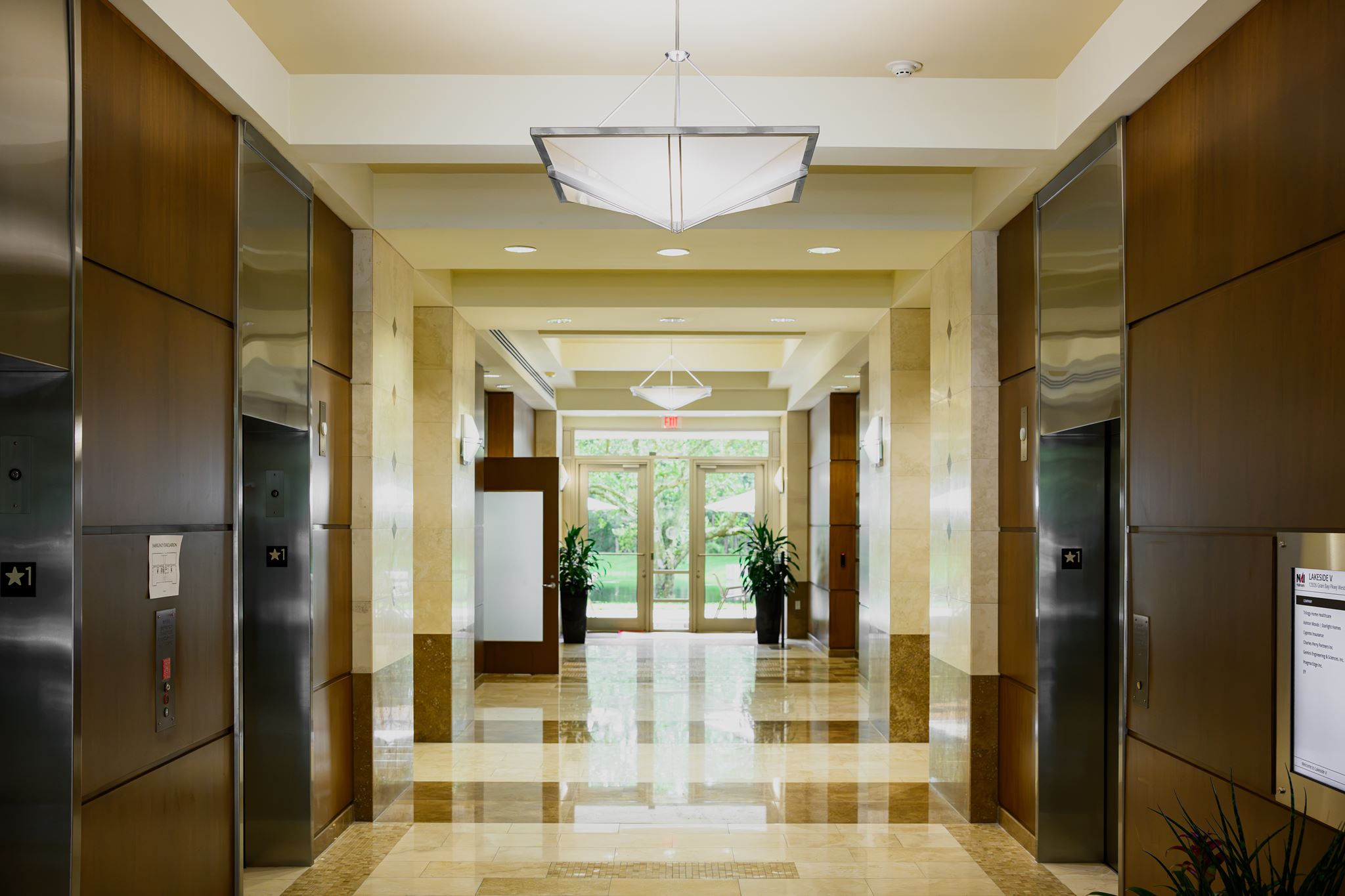 Modern building hallway with four elevator doors, shiny tiled floor, overhead lighting, potted plants, and large glass doors at the far end letting in natural light.