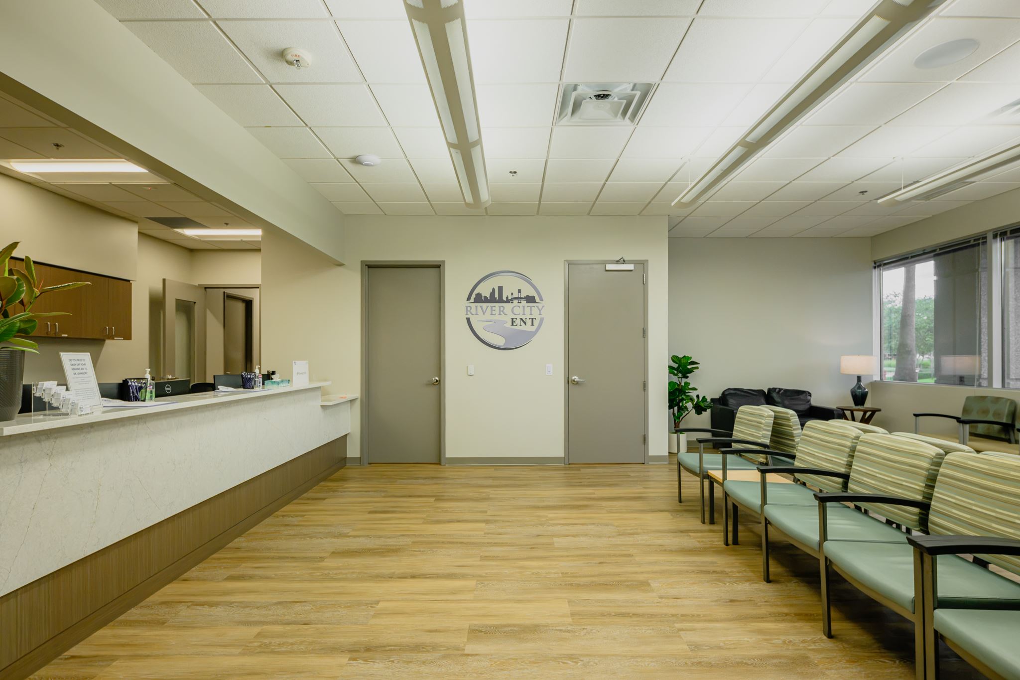 A medical office waiting room with rows of chairs, a reception desk on the left, and a sign reading “River City ENT” on the back wall.