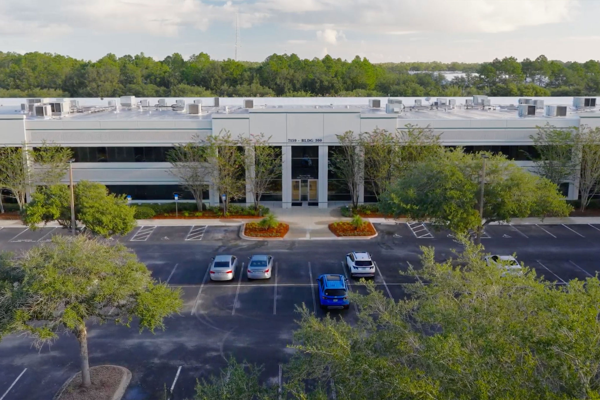 A two-story office building with large windows, several cars parked in front, landscaped greenery, and a backdrop of trees under a partly cloudy sky.